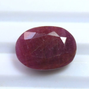 Puede incluir: Una gema de rubí facetada, de forma ovalada. La gema es de color rojo intenso con inclusiones internas visibles. El corte de la piedra crea múltiples superficies reflectantes, realzando su brillo. El rubí se presenta sobre un fondo blanco.