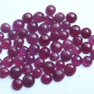May include: A selection of polished, round gemstones in various sizes. The gems are a deep, rich red colour, with a slight translucence. The stones are scattered across a white surface, creating a visually appealing display. These could be rubies.