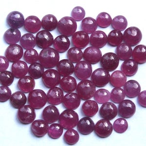 May include: A collection of polished, round gemstones in varying shades of red and purple. The gemstones are scattered across a white surface, showcasing their smooth, reflective surfaces. The sizes of the gemstones vary.