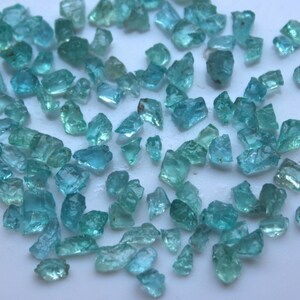 Pode incluir: Uma coleção de lascas de pedras preciosas azul turquesa. As lascas são de vários tamanhos e formas, algumas têm uma forma mais retangular e outras são mais irregulares. As lascas estão espalhadas sobre uma superfície branca.