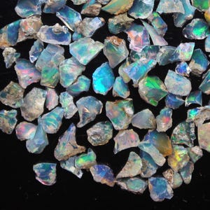 Puede incluir: Una colección de piedras preciosas de ópalo en bruto sin cortar. Los ópalos muestran una variedad de colores, incluyendo azules, verdes y destellos de tonos iridiscentes. Las piedras tienen forma irregular y parecen recién extraídas.