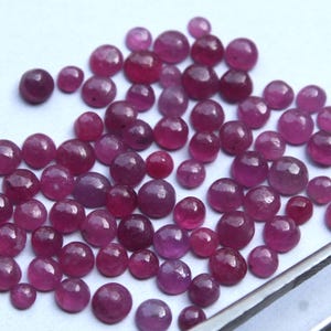 Könnte beinhalten: Eine Ansammlung kleiner, runder, polierter Rubin-Edelsteine in verschiedenen Größen. Die Edelsteine zeigen einen tiefroten bis violetten Farbton und sind auf einer weißen Oberfläche verstreut. Eine Pinzette ist in der unteren rechten Ecke sichtbar.