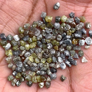 May include: A selection of raw diamonds in a variety of colours, including grey, yellow, and clear. The gemstones are small and irregularly shaped, with a natural, unpolished appearance. The diamonds are held in a hand, showcasing their raw beauty.