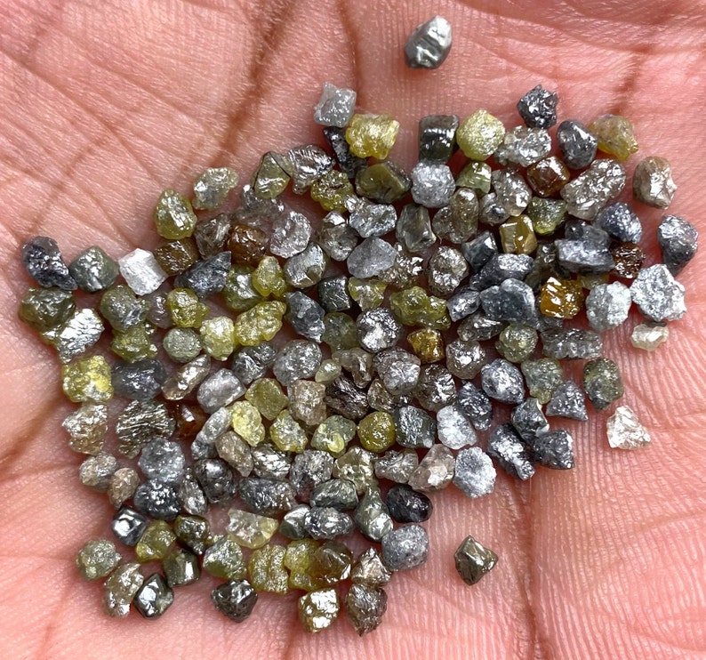 May include: A selection of raw diamonds in a variety of colours, including grey, yellow, and clear. The gemstones are small and irregularly shaped, with a natural, unpolished appearance. The diamonds are held in a hand, showcasing their raw beauty.