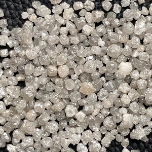 May include: A close-up view of a collection of small, rough, gray diamonds. The diamonds are scattered on a black surface.