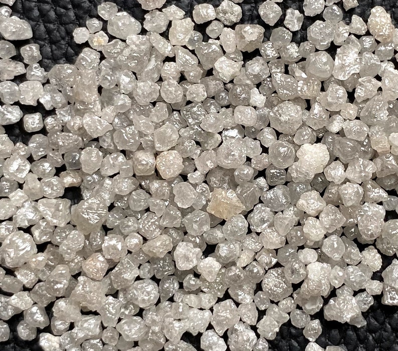 May include: A close-up view of a collection of small, rough, gray diamonds. The diamonds are scattered on a black surface.