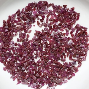 Op de afbeelding: Een close-up van een stapel kleine, ruwe, rode robijn edelstenen. De robijnen zijn verspreid over een wit oppervlak.