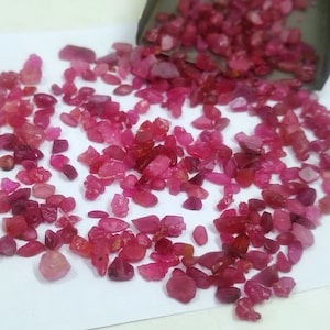May include: A collection of small, irregularly shaped, ruby-colored gemstones scattered on a white surface. The gemstones vary in shade from light pink to deep red, with a few still in a small, dark container.