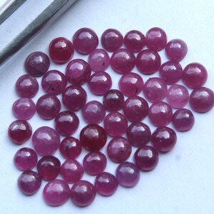 Könnte beinhalten: Eine Sammlung polierter, runder Rubin-Edelsteine in verschiedenen Rot- und Rosatönen. Die Edelsteine sind verstreut, mit einer teilweise sichtbaren Pinzette. Die Edelsteine haben eine glatte, reflektierende Oberfläche.