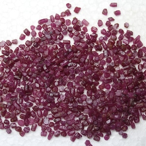 Pode incluir: Uma coleção de pequenas pedras preciosas de rubi em bruto. As pedras preciosas são de um vermelho profundo e rico, com variações de tom e forma. Estão espalhadas sobre uma superfície branca, criando uma aparência texturizada.