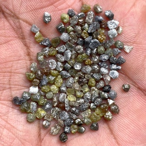May include: A collection of uncut diamonds in a range of colours, including grey, yellow, and clear. The gemstones are irregularly shaped and have a natural, unpolished appearance. The diamonds are small, likely under 1 carat each.