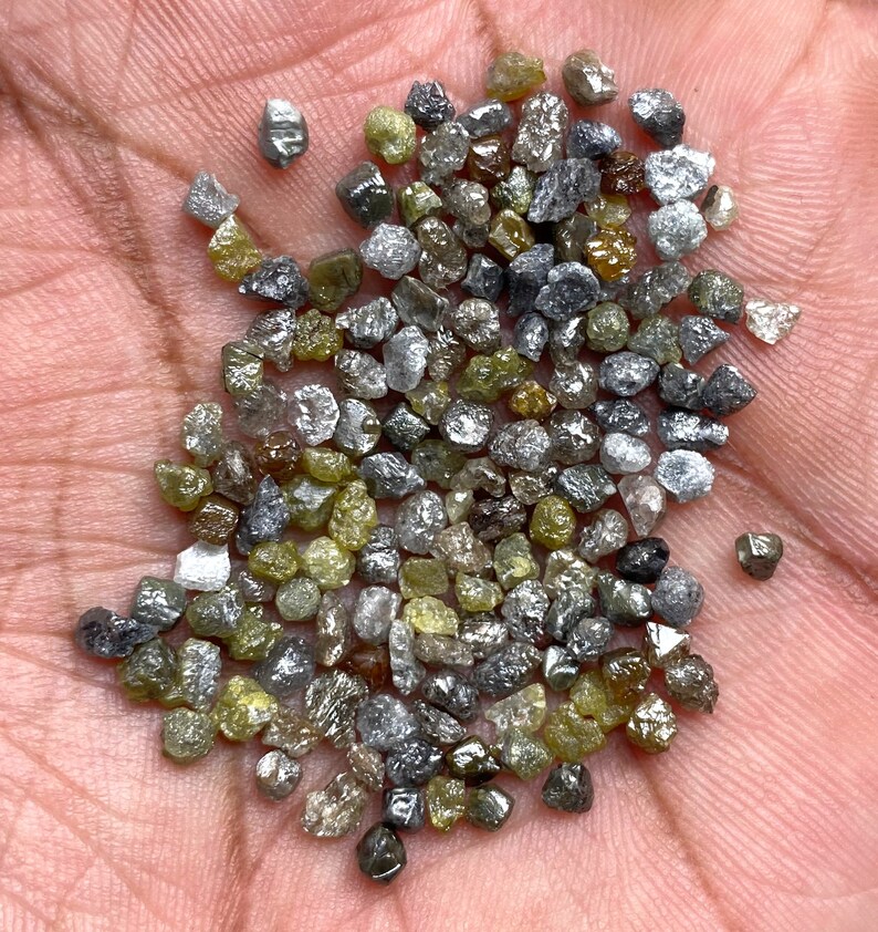 May include: A collection of uncut diamonds in a range of colours, including grey, yellow, and clear. The gemstones are irregularly shaped and have a natural, unpolished appearance. The diamonds are small, likely under 1 carat each.