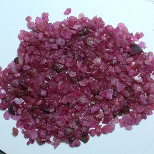 K&ouml;nnte beinhalten: Ein Haufen roher, ungeschliffener Rubin-Edelsteine. Die Rubine haben eine tiefe, satte rote Farbe und weisen unterschiedliche Formen und Gr&ouml;&szlig;en auf. Die Edelsteine sind auf einer wei&szlig;en Oberfl&auml;che verstreut und zeigen ihren nat&uuml;rlichen, unpolierten Zustand.