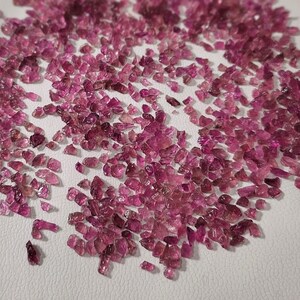 May include: A large quantity of pink tourmaline rough gemstones. The rough stones are irregularly shaped and have a variety of sizes. The stones are scattered on a white surface.