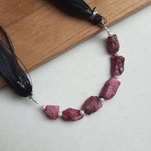 May include: A strand of pink ruby gemstone beads with white pearl accents, strung on a silver wire with black tassel ends.