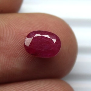 Peut inclure: Pierre précieuse de forme ovale, un rubis d'un rouge profond. La gemme présente de multiples facettes qui reflètent la lumière, créant un effet scintillant. Le rubis est tenu entre deux doigts, mettant en valeur sa taille et sa couleur.