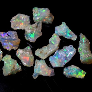 Puede incluir: Una colección de piedras preciosas de ópalo en bruto de diferentes formas y tamaños. Los ópalos son iridiscentes con destellos de verde, azul y rojo.