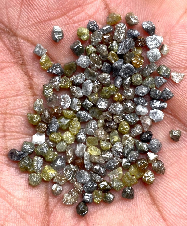 May include: A scattering of raw diamonds in shades of grey, yellow-green, and clear. The gemstones are small and irregularly shaped, with a natural, unpolished appearance. The diamonds are held in a hand, highlighting their natural beauty.