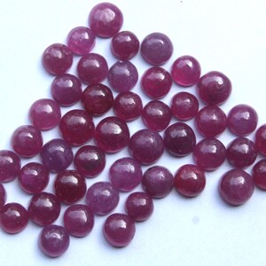 May include: A collection of round, polished ruby gemstones in varying shades of red and purple. The gemstones are scattered across a white surface, showcasing their smooth, reflective surfaces. Ideal for jewelry making.