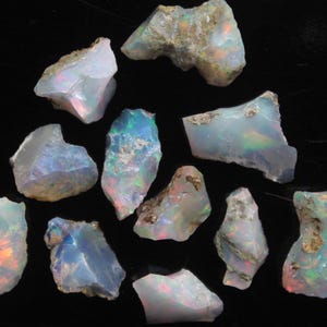 Puede incluir: Una colección de piedras preciosas de ópalo en bruto, que muestran una variedad de formas y tamaños. Los ópalos exhiben un color base blanco lechoso con destellos iridiscentes de azul, verde y rojo. Se aprecia la textura rugosa y las formas naturales.