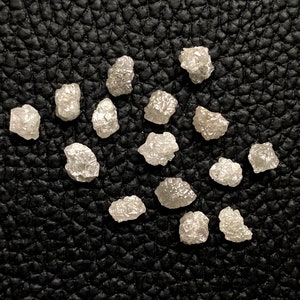 Op de afbeelding: Een verzameling ruwe, ongeslepen diamanten, elk met een unieke, onregelmatige vorm. De diamanten hebben een bleke, doorschijnende kleur en liggen op een zwarte, getextureerde ondergrond. De afbeelding is goed belicht, waardoor de facetten en de natuurlijke schoonheid van de edelstenen worden benadrukt.