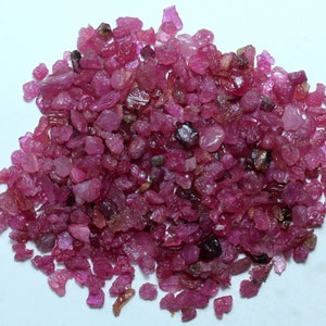 May include: A pile of rough-cut ruby gemstones. The stones vary in size and shape, displaying a range of pink and red hues. Some stones have a translucent quality, while others are more opaque. The gemstones are scattered on a white surface.