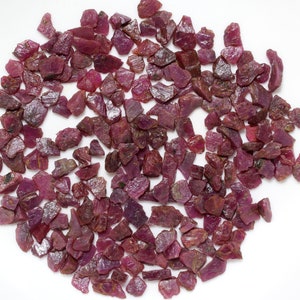 Könnte beinhalten: Eine Ansammlung von ungeschliffenen Rubin-Edelsteinen in verschiedenen Größen und Formen. Die Edelsteine zeigen einen tiefroten Farbton mit einigen dunkleren Bereichen. Die rohen Rubinkristalle sind auf einer weißen Oberfläche verstreut und zeigen ihren natürlichen, unpolierten Zustand.