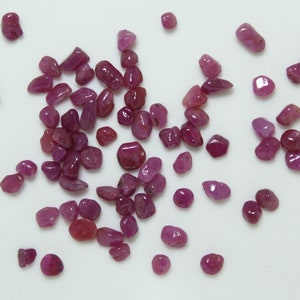 May include: A collection of small, tumbled ruby gemstones in varying shapes and sizes. The gemstones display a range of deep red and pink hues, scattered on a white surface. These natural stones are suitable for jewelry making or collecting.