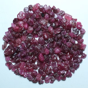 May include: A large pile of small, rough, pink ruby gemstones. The rubies are various shades of pink and have a natural, unpolished look.
