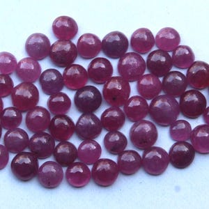Könnte beinhalten: Eine Sammlung runder, polierter Edelsteine in verschiedenen Rot- und Lilatönen. Die Edelsteine sind auf einer hellen Oberfläche verstreut und zeigen ihre glatten, reflektierenden Oberflächen. Die Steine scheinen Cabochons zu sein.