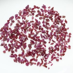 Könnte beinhalten: Eine große Sammlung kleiner, roher, roter Rubin-Edelsteine. Die Rubine sind auf einer weißen Oberfläche verstreut.