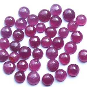 Könnte beinhalten: Eine Sammlung runder, polierter Rubin-Edelsteine in verschiedenen Größen. Die Edelsteine haben eine tiefe, satte rote Farbe mit einer glatten, reflektierenden Oberfläche. Die Edelsteine sind auf weißem Hintergrund verstreut.