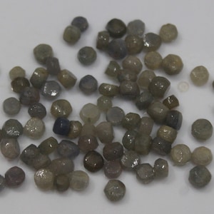 May include: A collection of small, irregularly shaped gemstones in various shades of gray, brown, and blue. The gemstones are scattered across a white surface, with a mix of rounded and faceted forms. These could be used for jewelry making.