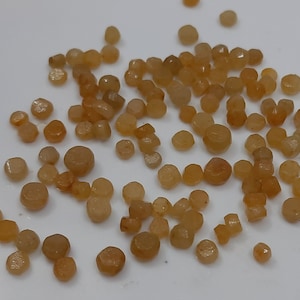 May include: A collection of small, light brown, faceted gemstones. The gemstones are scattered on a white surface.