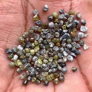 May include: A collection of uncut diamonds in a variety of colours, including clear, grey, yellow, and brown. The gemstones are irregularly shaped and have a natural, unpolished appearance. The diamonds are small, with a rough texture.