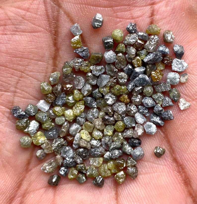 May include: A collection of uncut diamonds in a variety of colours, including clear, grey, yellow, and brown. The gemstones are irregularly shaped and have a natural, unpolished appearance. The diamonds are small, with a rough texture.