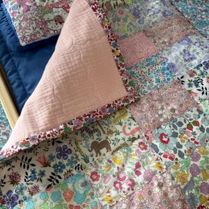 May include: A patchwork quilt and pillow set with a floral design in pink, blue, and white. The quilt has a pink backing and a patterned border. The pillow rests on a blue surface, and the quilt is on a wooden frame. The quilt is made of several patches.