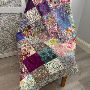 May include: A colourful patchwork quilt draped over a wooden chair. The quilt is made from squares of floral and patterned fabrics in pinks, purples, blues, and greens. The edges are finished with a black and white floral border. A vibrant textile.