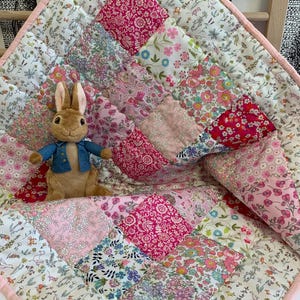 May include: A patchwork quilt featuring squares of floral fabric in pink and white, with a light pink border. A stuffed bunny in a blue jacket rests on the quilt. The quilt's design incorporates various floral patterns in shades of pink and red.