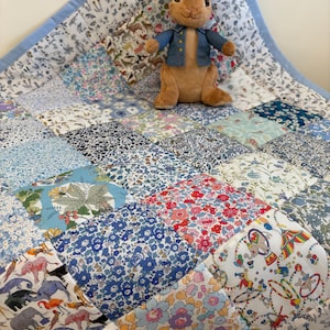 May include: A patchwork quilt featuring a mix of floral and animal print fabrics in blue, pink, and white. A stuffed rabbit wearing a blue jacket is placed on the quilt. The quilt has a light blue border.