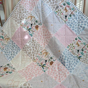 May include: A patchwork quilt with a mix of patterns and colours. The quilt features squares of pink, blue, and beige fabrics with giraffe, lion, and floral designs. The border is striped.