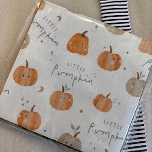 May include: A square fabric item wrapped in clear plastic featuring a repeating pattern of orange pumpkins with faces, small stars, and crescent moons on a light background. The text Little Pumpkin is printed in black script. A black and white striped ribbon.