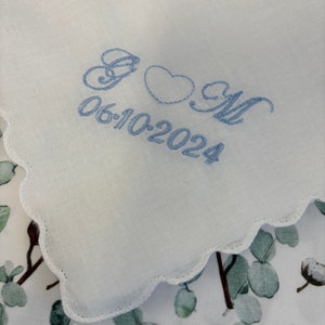 May include: White handkerchief with a scalloped edge, embroidered with the initials "G & M", a heart, and the date "06-10-2024" in light blue thread. The handkerchief is displayed on a floral patterned fabric.