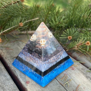May include: A clear crystal pyramid with layers of black, white, and blue resin. The pyramid is filled with small white skulls, black stones, and copper wire.