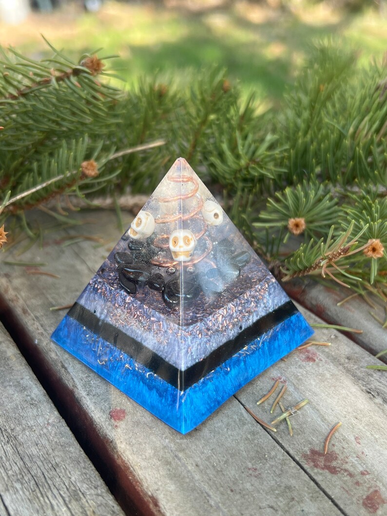 May include: A clear crystal pyramid with layers of black, white, and blue resin. The pyramid is filled with small white skulls, black stones, and copper wire.