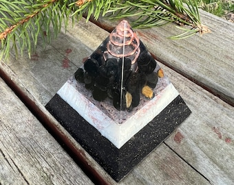 May include: A black, white, and gold pyramid with a copper wire spiral inside. The pyramid is filled with black stones and has a white base.