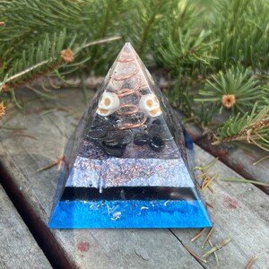 May include: A blue, black, and white crystal pyramid with a copper wire spiral and two white skull beads. The pyramid is sitting on a wooden surface.