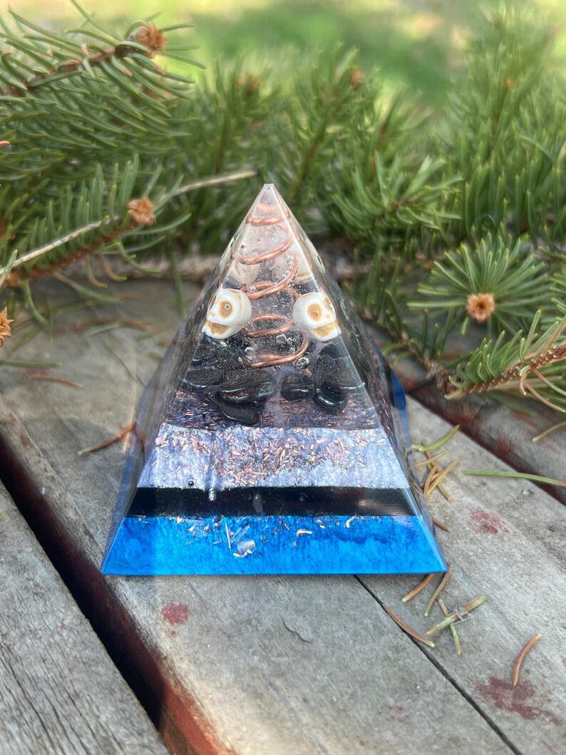 May include: A blue, black, and white crystal pyramid with a copper wire spiral and two white skull beads. The pyramid is sitting on a wooden surface.