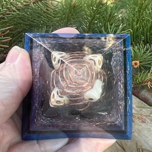 May include: A clear crystal pyramid with a copper wire coil inside. The pyramid is surrounded by four white crystal orbs with gold accents. The pyramid is set on a blue base with a glittery finish.