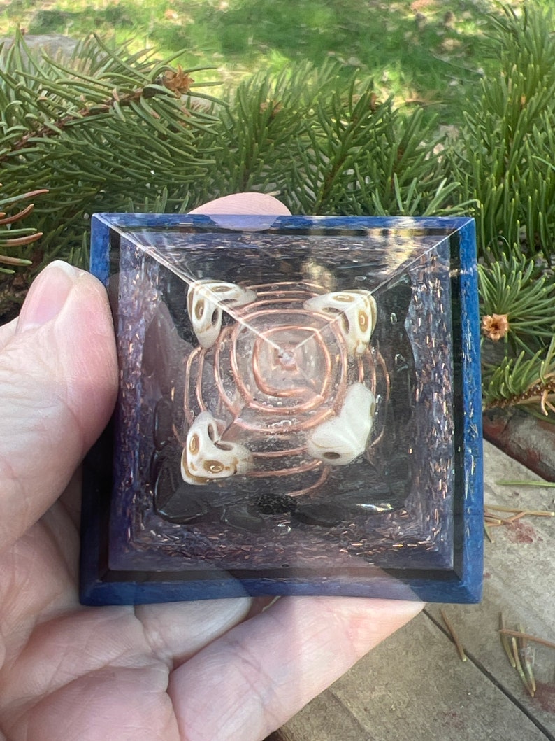 May include: A clear crystal pyramid with a copper wire coil inside. The pyramid is surrounded by four white crystal orbs with gold accents. The pyramid is set on a blue base with a glittery finish.